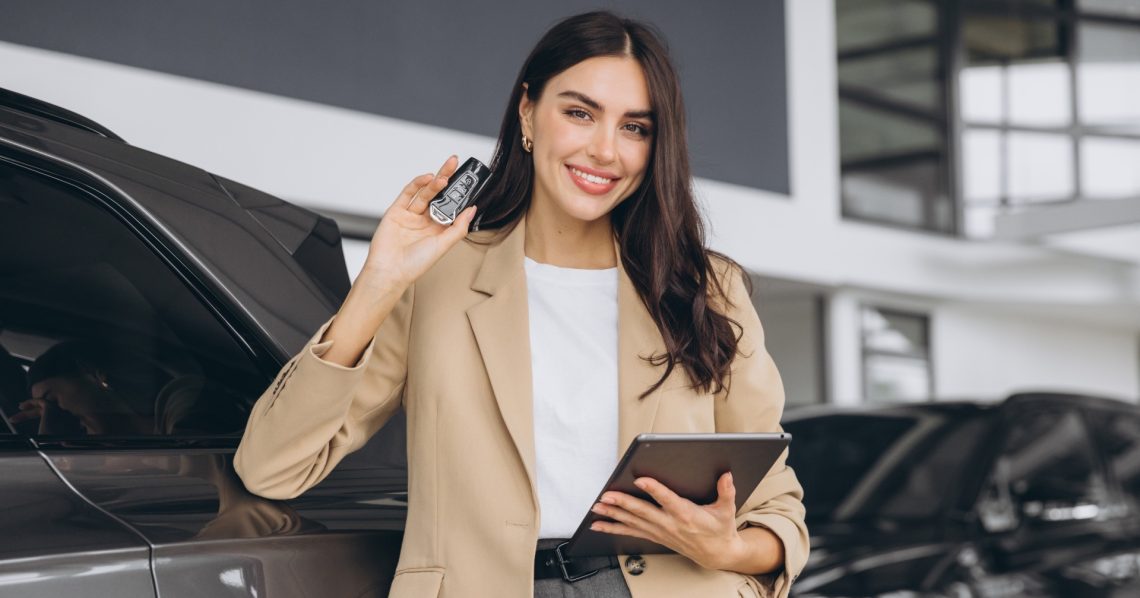 Beautiful smiling saleswoman in full suit in dealership on cars background with car keys and tablet in hands